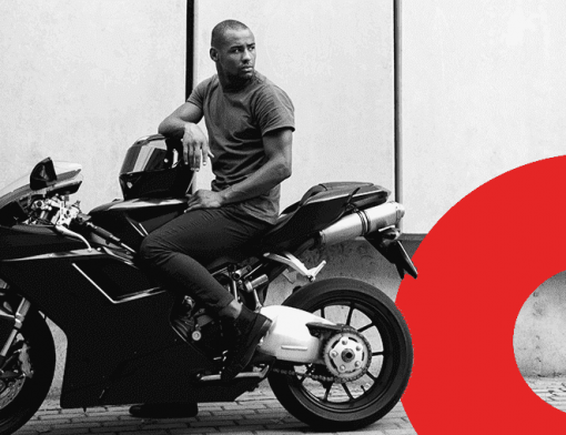 Capa de artigo isenção de ipva para motos | DOK Despachante | Descrição: Motociclista jovem negro segura o capacete em uma mão, montado sobre a moto. Imagem em preto e branco.