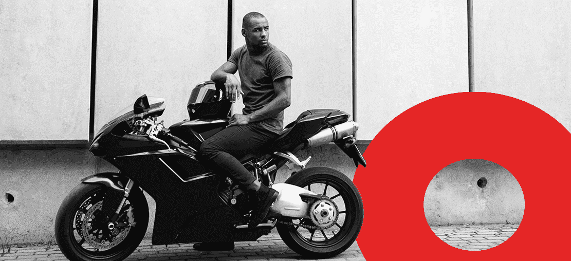 Capa de artigo isenção de ipva para motos | DOK Despachante | Descrição: Motociclista jovem negro segura o capacete em uma mão, montado sobre a moto. Imagem em preto e branco.
