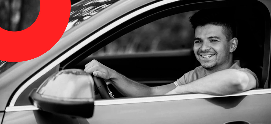Capa de artigo Bradesco Seguros - DOK Despachante; Descrição: foto em preto e branco, jovem motorista homem e branco sorri ao segurar volante.
