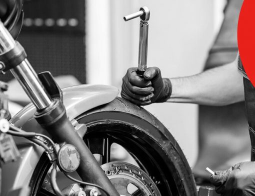 Capa de artigo sobre acessórios para motos | DOK Despachante; Descrição: imagem em preto e branco de pessoa de luvas segurando uma chave de roda ao lado de roda dianteira de moto. Imagem em preto e branco.